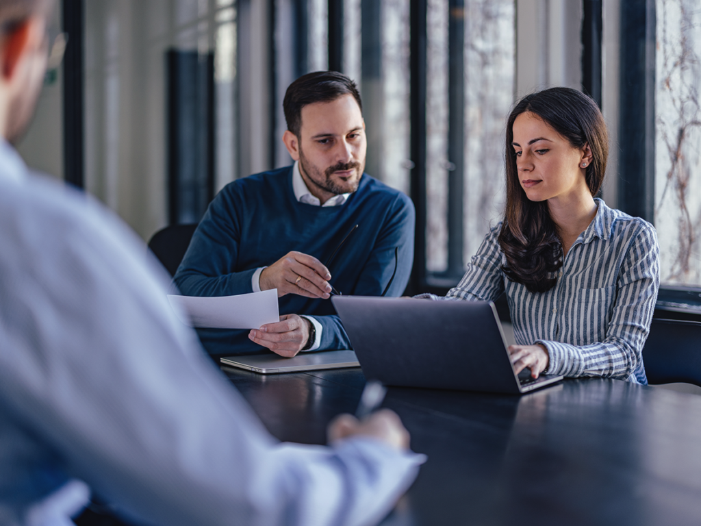 Two employees at a table discussing a laptop screen focused on reactive vs proactive management in operations.